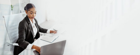 Woman Working on Laptop and Taking Notes at Desk in Office Setting During Daytimeの写真素材