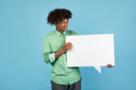African american teenager holding blank speech bubble and looking at it, standing over blue studio...