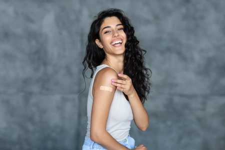 Lovely young woman getting vaccinated against covid-19, showing arm with band aid on grey studio backgroundの写真素材