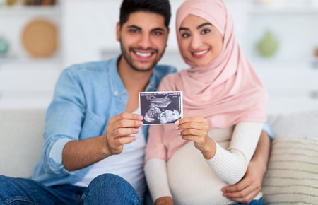 Happy arab family demonstrating ultrasound scan and smiling to camera, sitting on sofa at home
