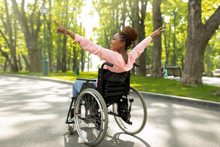 Handicapped black woman in wheelchair on walk at city park, showing victory gesture, feeling positive and motivatedの写真素材