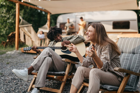 Happy black guy playing guitar, his female friend singing, sitting in lounge chairs near RV, camping on summer vacationの写真素材