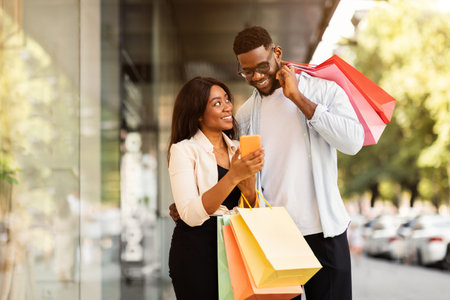 Portrait of happy black couple using phone with shopping bagsの写真素材