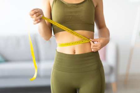 Cropped view of slim Indian woman measuring her waist with tape measure at home, closeup