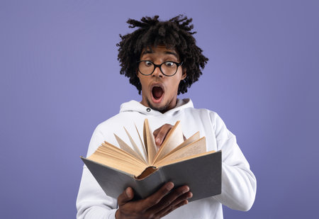 Black teen student looking at textbook in shock, afraid of too much homework on violet studio...