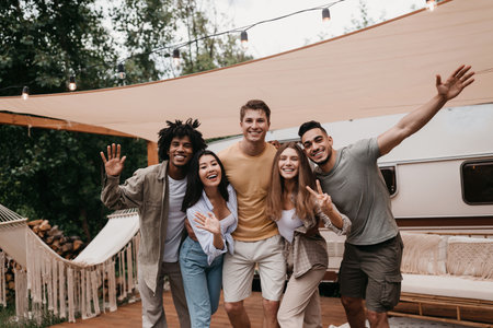 Portrait of cheery young international friends posing near camper van, smiling and waving at camera,...