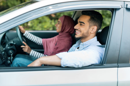 Happy cheerful young muslim woman in hijab is driving car, husband enjoying journey with open windowの写真素材