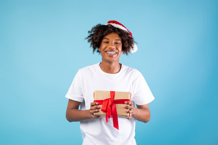 Christmas suprise. Joyful black guy in santa hat holding gift box, posing over blue background and smiling at cameraの写真素材