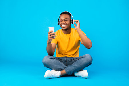 African Guy Listening Music Using Smartphone Wearing Headphones, Blue Background
