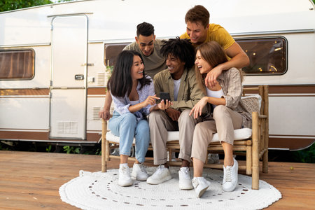Young multinational friends using smartphone near RV outdoors, having online video call, enjoying autumn camping tripの写真素材