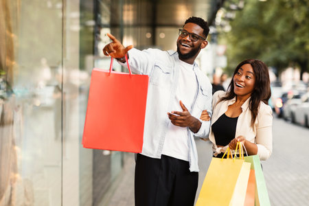Excited black couple with shopping bags pointing at windowの写真素材