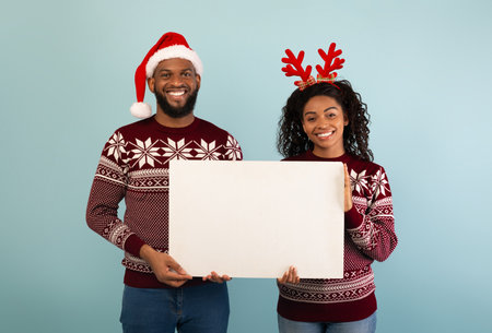 Positive black couple in Christmas outfits holding white poster with empty space, standing over blue backgroundの写真素材