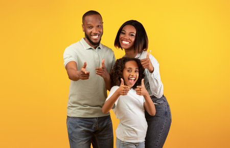 Hey, you. Excited black parents and their daughter pointing fingers at camera, standing over yellow backgroundの写真素材