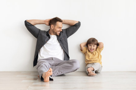 Relax time. Happy little boy sitting with daddy and laughing to camera, leaning wall with hands behind headの写真素材