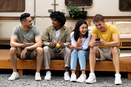 Diverse friends sitting near motorhome, talking to each other, using smartphone, spending time...