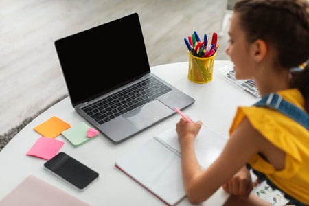 Girl sitting at desk, using blank laptop, writing in textbook