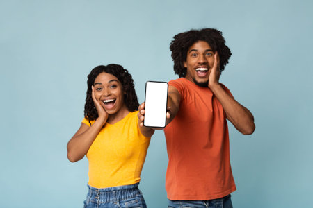 Amazed african-american couple showing smartphone with blank screen, mockupの写真素材