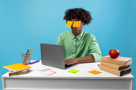 Exhausted teen guy with painted eyes on stickers sitting at desk using laptop over blue background
