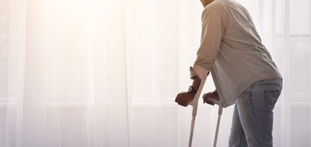 Man Using Crutches Looking out the Window in a Bright Room During the Dayの写真素材