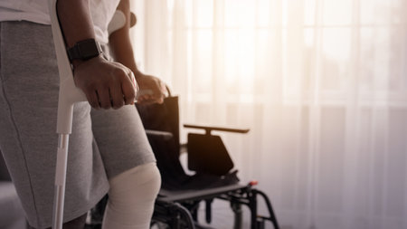 Man With Dark Skin Uses Crutches While Standing Beside a Wheelchair in a Bright Room
