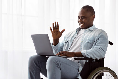 Man Using a Wheelchair Smiles and Waves While Video Chatting on a Laptop Indoors