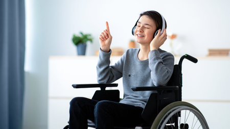 Young Boy With Light Skin Using a Wheelchair Enjoys Music With Headphones at Homeの写真素材