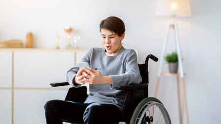 Young Man Using a Wheelchair Enjoys Time on Smartphone While Sitting Indoorsの写真素材