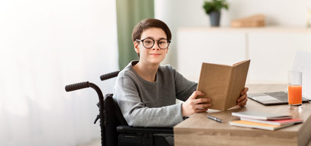 Boy With Glasses Using a Wheelchair Reads a Book at a Desk in a Bright Roomの写真素材