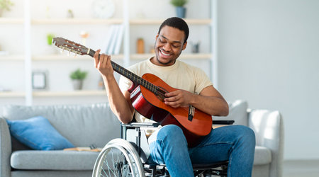 Young Man Using a Wheelchair Plays Guitar at Home During Daytime With a Smileの写真素材