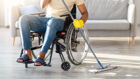 Man Using a Wheelchair Mops the Floor in a Bright Living Room With a Gray Couchの写真素材