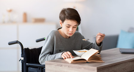 Young Boy Using a Wheelchair Enjoys Breakfast While Reading a Book in a Cozy Indoor Settingの写真素材