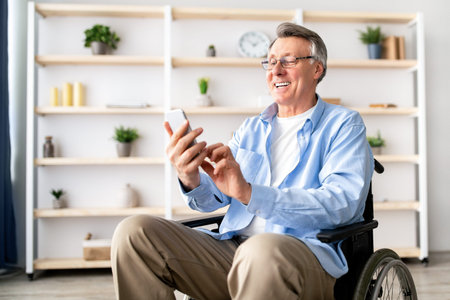 Older Man Using Smartphone While Sitting in a Wheelchair in a Bright Room With Shelvesの写真素材