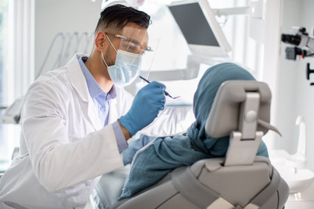 Middle-Eastern Male Dentist In Medical Mask And Face Shield Checking Patients Teethの写真素材