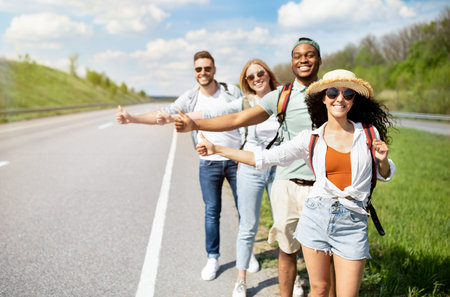 Young multiracial friends standing on highway, showing hitchhiking gesture, stopping car, going on journey togetherの写真素材