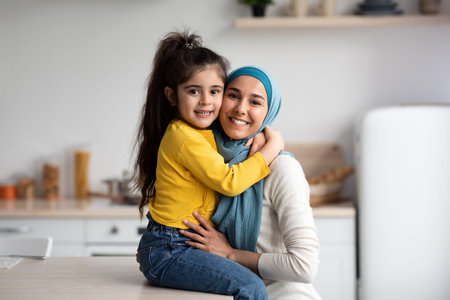 Happy Muslim Mother In Hijab And Little Daughter Posing In Kitchen Interiorの写真素材