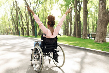 Full length of black woman in wheelchair enjoying walk in city park, showing peace gesture with both hands, back viewの写真素材