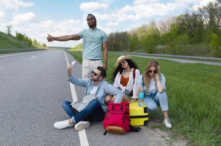 Young multiracial friends hitchhiking on highway, trying to stop car without success, feeling tired and upset on road tripの写真素材