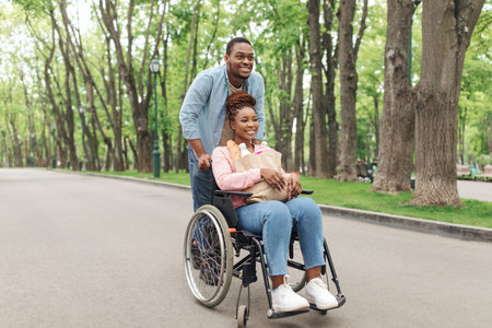 Cheerful young black woman in wheelchair carrying bag with products, going shopping with her...