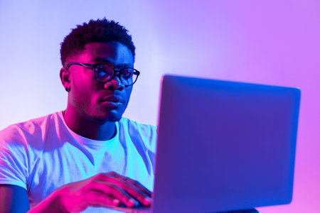 Focused Young Black Man Working on Laptop in Neon Light During Online Meeting