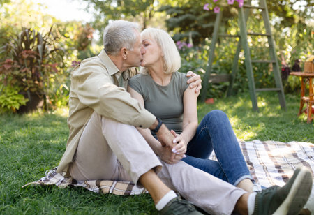 Married senior couple resting outdoors and kissing, enjoying holiday while sitting on blanket on grass in gardenの写真素材