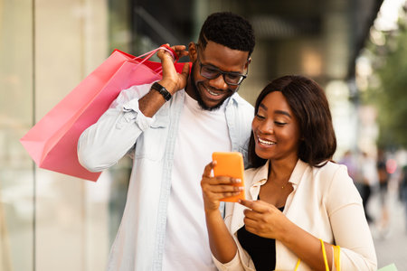 Happy black couple using phone with shopping bagsの写真素材