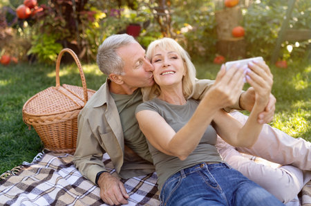 Happy senior couple taking selfie on smartphone, having picnic and resting in garden outside, enjoying autumn natureの写真素材