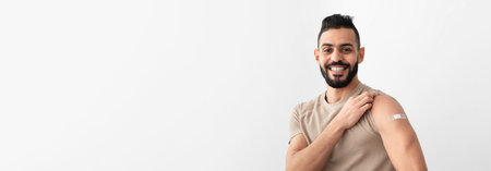 Cheerful Arab man showing arm with band aid after receiving covid vaccine injection, smiling,...