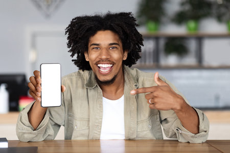 Glad happy millennial african american curly man pointing finger at smartphone with blank screen