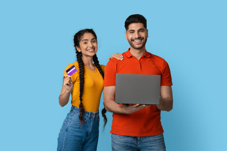 Smiling Young Couple Holding a Laptop and a Card Against a Blue Background