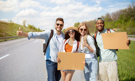Portrait of happy diverse friends hitchhiking together on highway, making thumb up gestures, holding empty signsの写真素材