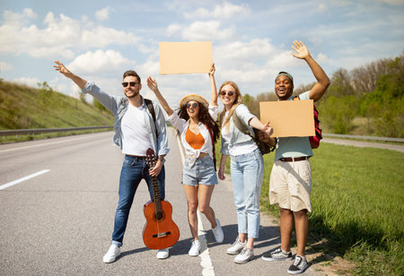 Group of multiracial friends with guitar and empty sign hitchhiking on road, waving down car, traveling by autostopの写真素材