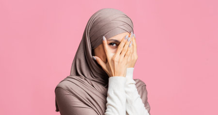 Portrait of shy modest islamic woman in hijab peeking through fingersの写真素材