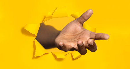 Hand stretching through hole in torn yellow paper, offering helping handの写真素材