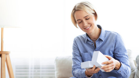 Woman Smiles While Opening a Gift Box in a Cozy Indoor Setting During Daytimeの写真素材